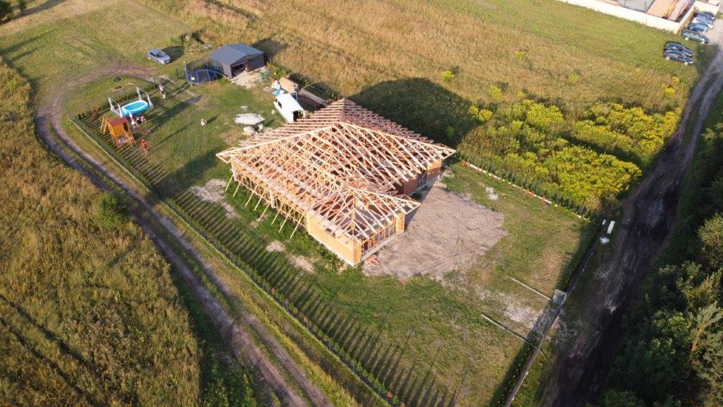 Widok z lotu ptaka na budowę domu jednorodzinnego z widoczną konstrukcją drewnianą dachu, ceglanymi ścianami i otaczającym terenem z elementami rekreacyjnymi.