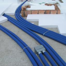 F.H.P. Prodmax - Niebieskie, karbowane rury wentylacyjne prowadzone w warstwie izolacji z białego styropianu, z widoczną skrzynką rozdzielczą i fragmentami ceglanej konstrukcji.