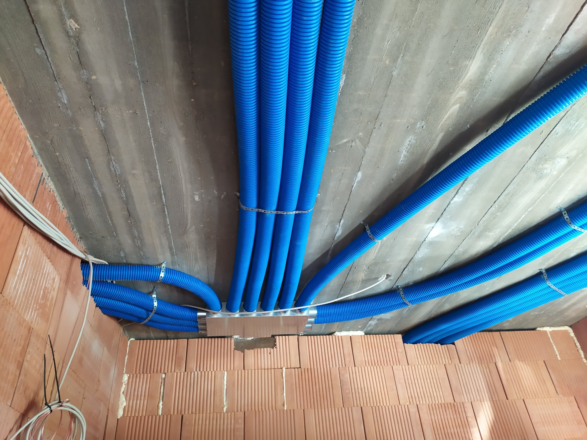 Niebieskie, karbowane rury wentylacyjne przymocowane do sufitu z betonu architektonicznego, widoczne także przewody elektryczne i fragment ściany z cegieł.