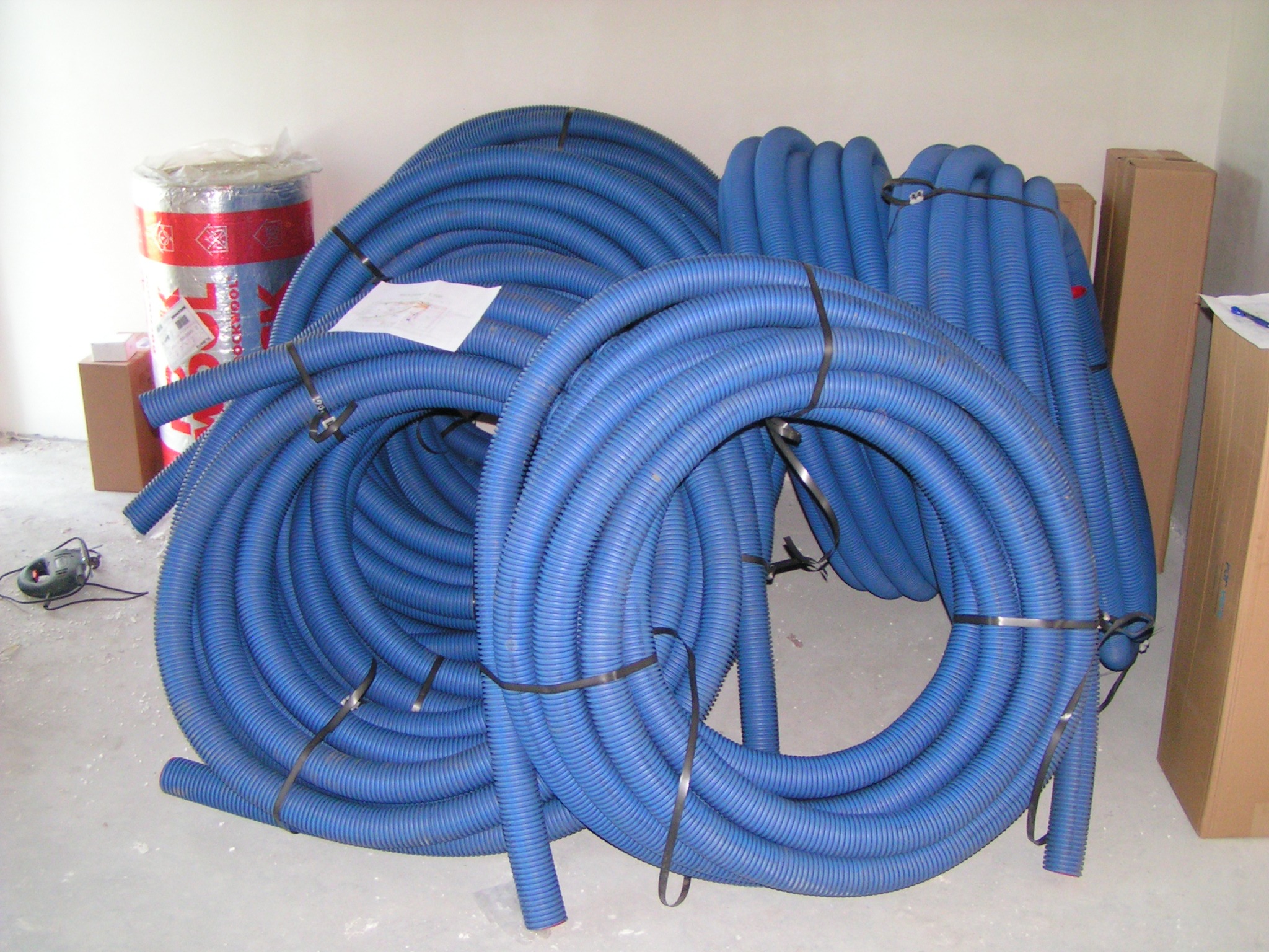 Stos niebieskich, karbowanych przewodów wentylacyjnych zwiniętych w kręgi, zabezpieczonych czarnymi opaskami zaciskowymi, obok rolka wełny mineralnej i kartony, na szarym betonowym podłożu.