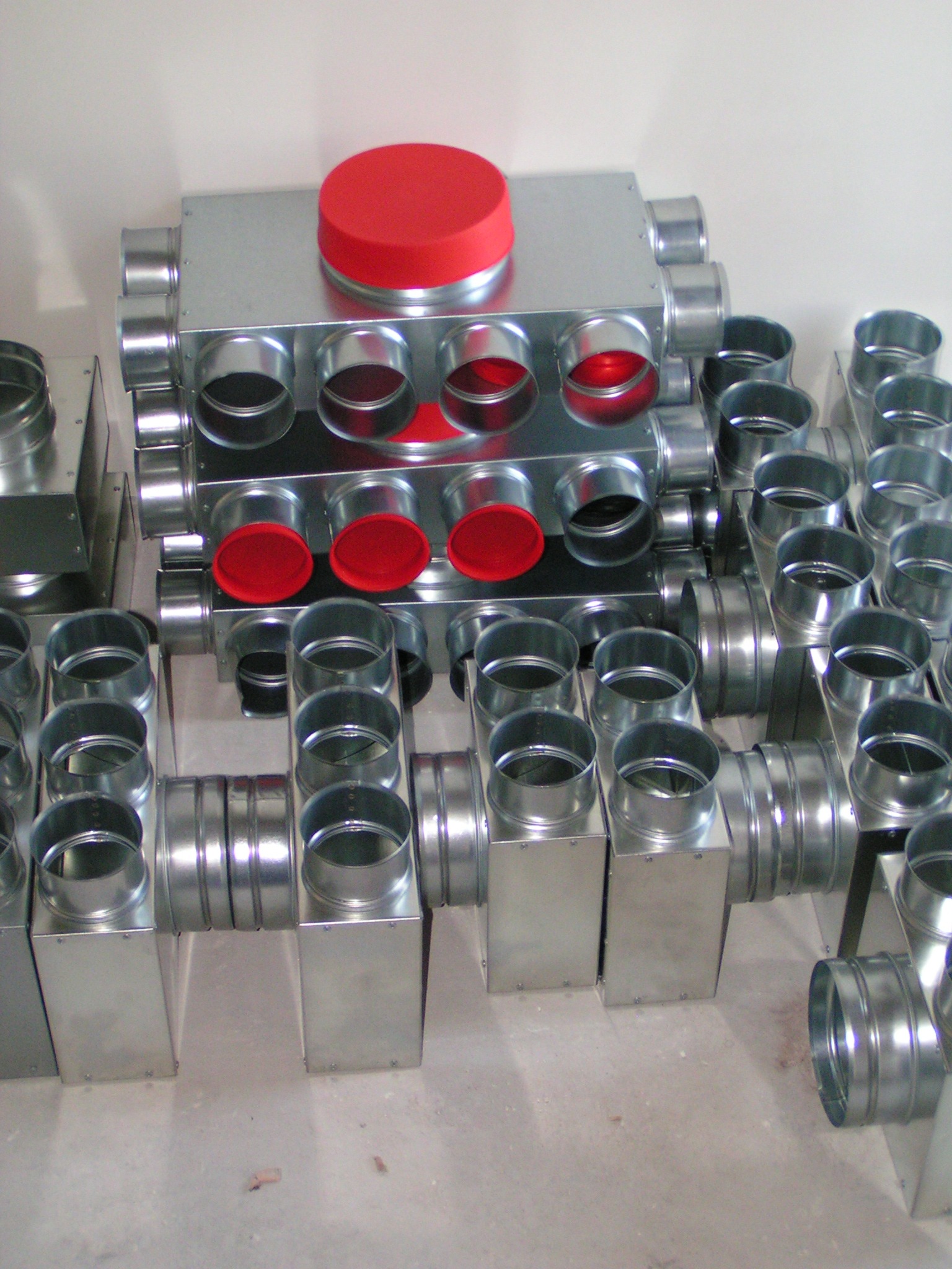 Stos metalowych skrzynek wentylacyjnych z okrągłymi otworami i czerwonymi zaślepkami, ułożonych na podłodze w magazynie.