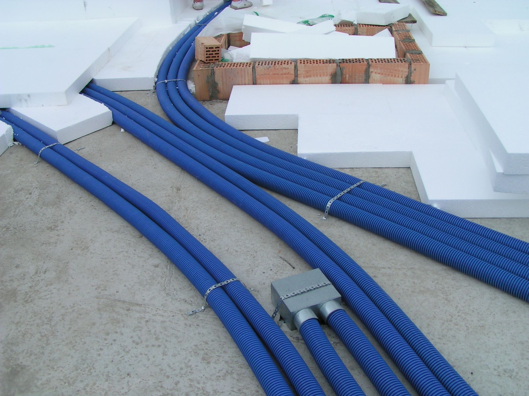 Niebieskie, karbowane rury wentylacyjne prowadzone w warstwie izolacji z białego styropianu, z widoczną skrzynką rozdzielczą i fragmentami ceglanej konstrukcji.