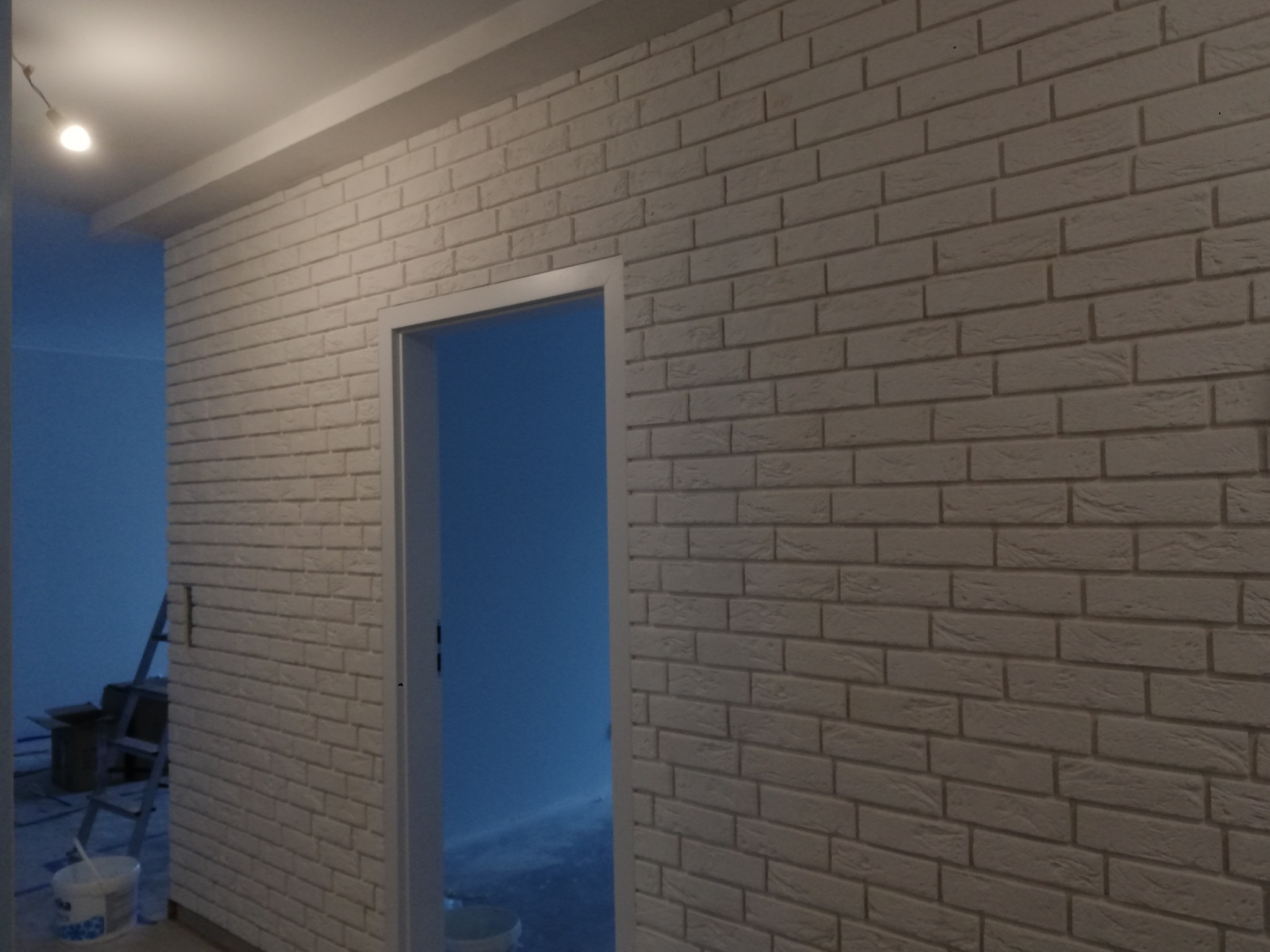Ściana wewnętrzna wykończona dekoracyjną cegłą w odcieniach bieli, widoczna rama drzwi i fragment pomieszczenia w trakcie remontu z drabiną i wiadrem farby w tle.