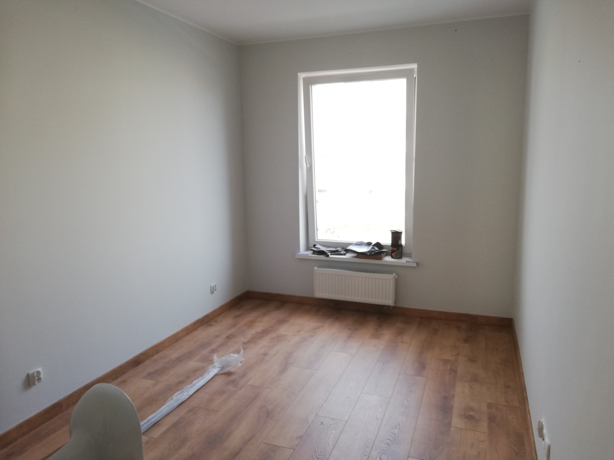 Pomalowane na szaro puste pomieszczenie z drewnianą podłogą, grzejnikiem pod oknem i elementami w folii na podłodze, gotowe do dalszych prac wykończeniowych.