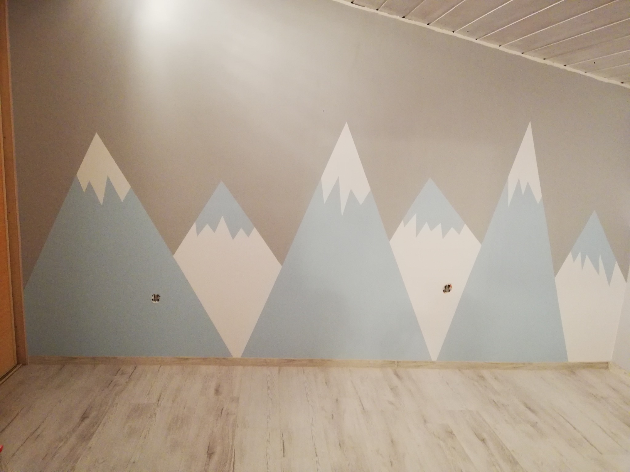 Ściana pokoju dziecięcego z ręcznie malowanym motywem gór w odcieniach szarości, błękitu i bieli, widoczne gniazdka elektryczne na ścianie, podłoga laminowana imitująca drewno.