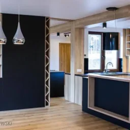 Nowoczesna kuchnia z czarnymi frontami i drewnianymi akcentami, wyspa z blatem, wiszące lampy w kształcie kropli, drewniane belki stropowe i logo firmy w rogu.