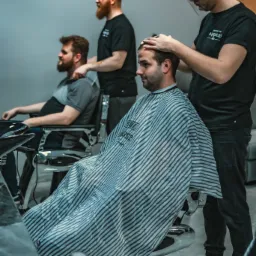 Wnętrze salonu z trzema mężczyznami: klient w pelerynie w paski podczas strzyżenia, drugi klient czeka w fotelu, trzeci w tle. Na ścianie widoczny zegar.