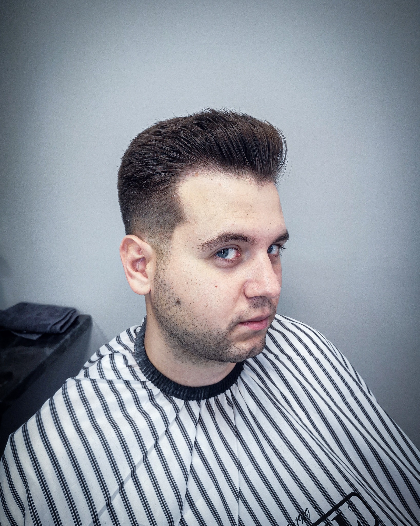 Mężczyzna z nową, starannie wystylizowaną fryzurą typu pompadour, ubrany w pelerynę fryzjerską w czarno-białe paski, spogląda w bok.