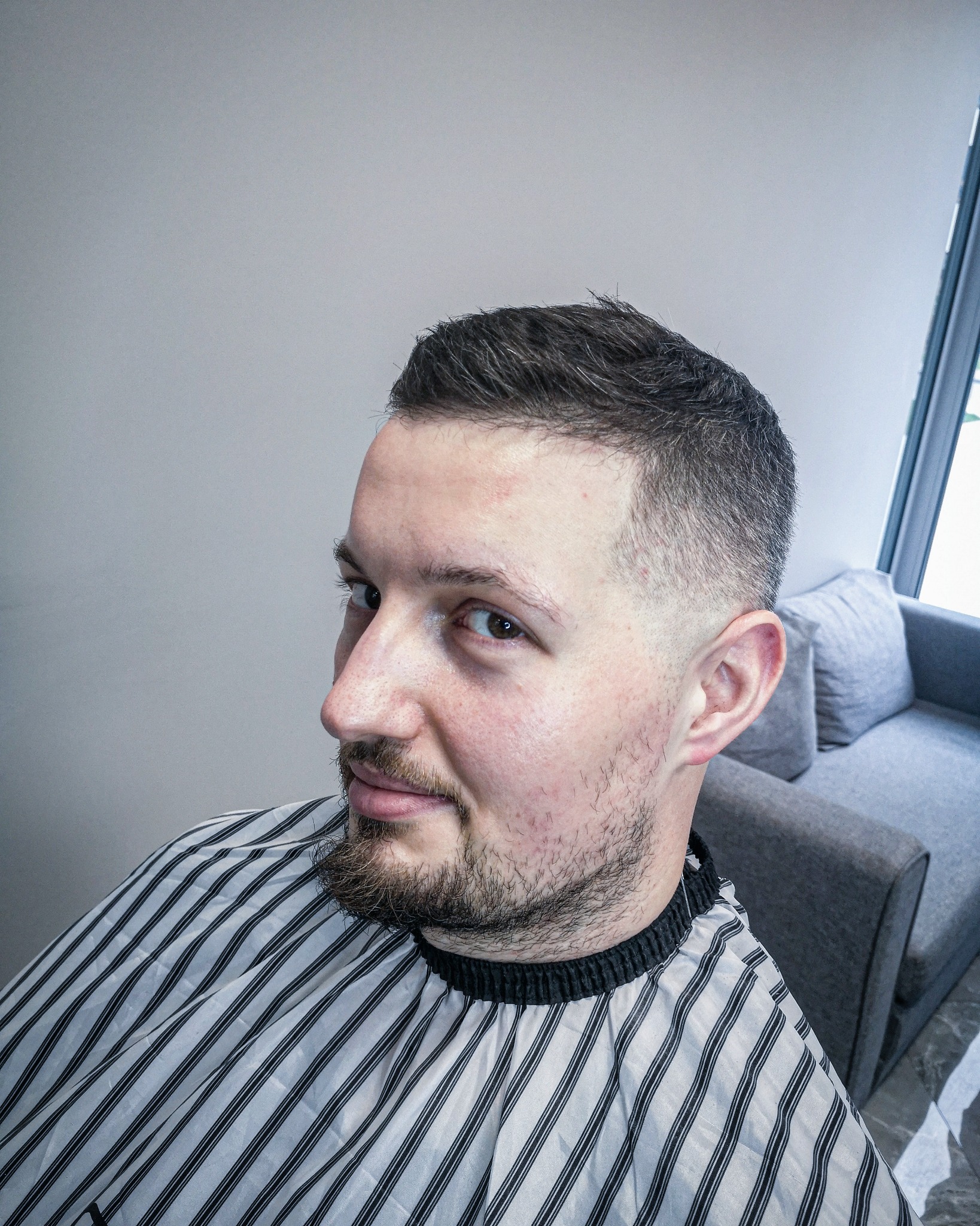 Mężczyzna z krótką, modną fryzurą i delikatnym zarostem, ubrany w pelerynę fryzjerską w czarno-białe paski, spogląda w bok z lekkim uśmiechem.
