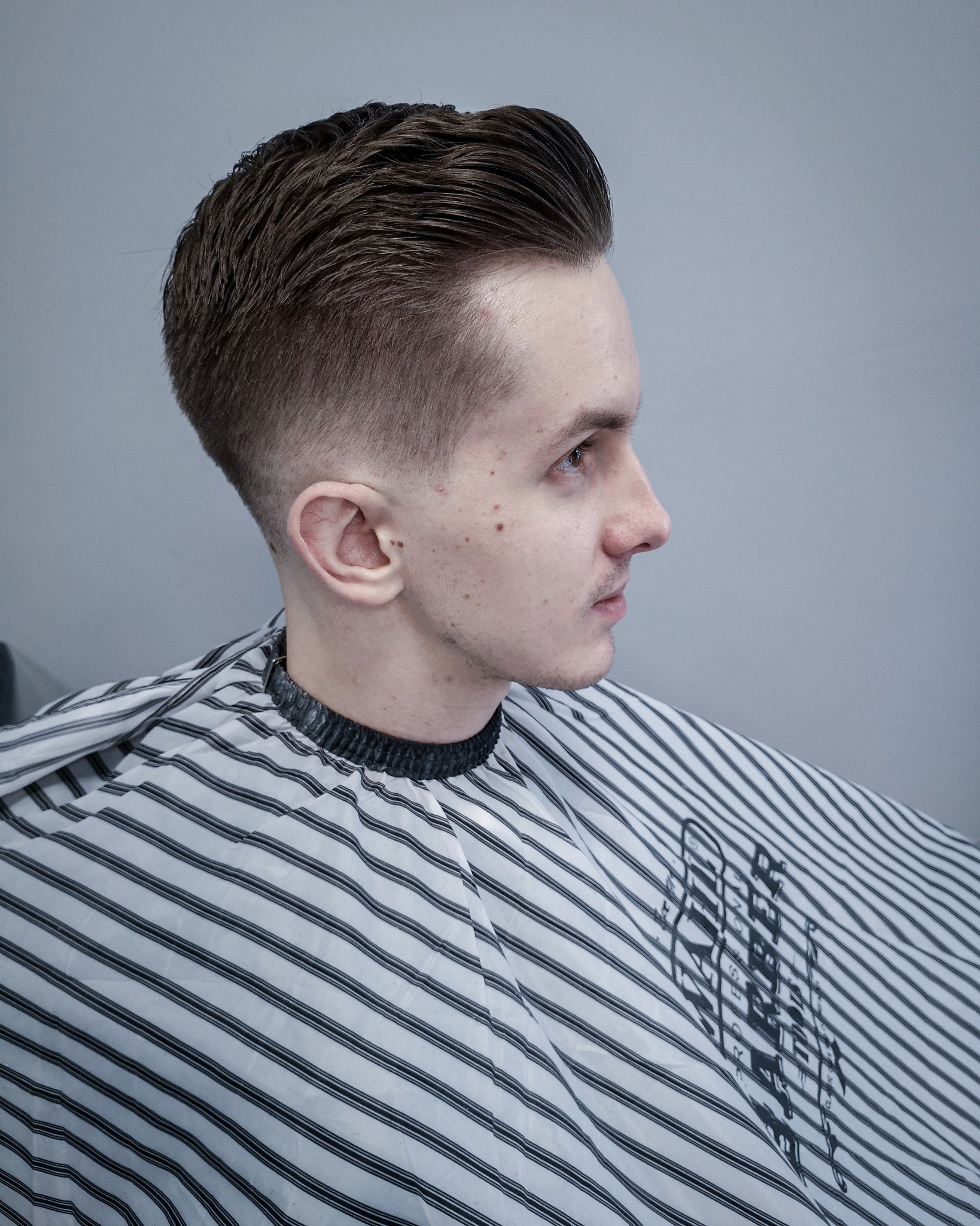 Młody mężczyzna z modną fryzurą typu slick back, ubrany w pelerynę w czarno-białe paski, patrzy w prawo, widoczne detale cieniowania i zaczesania włosów.