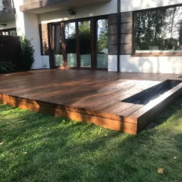 Nowoczesny, drewniany taras przylegający do domu z wbudowaną donicą, widok z poziomu trawnika w słoneczny dzień.