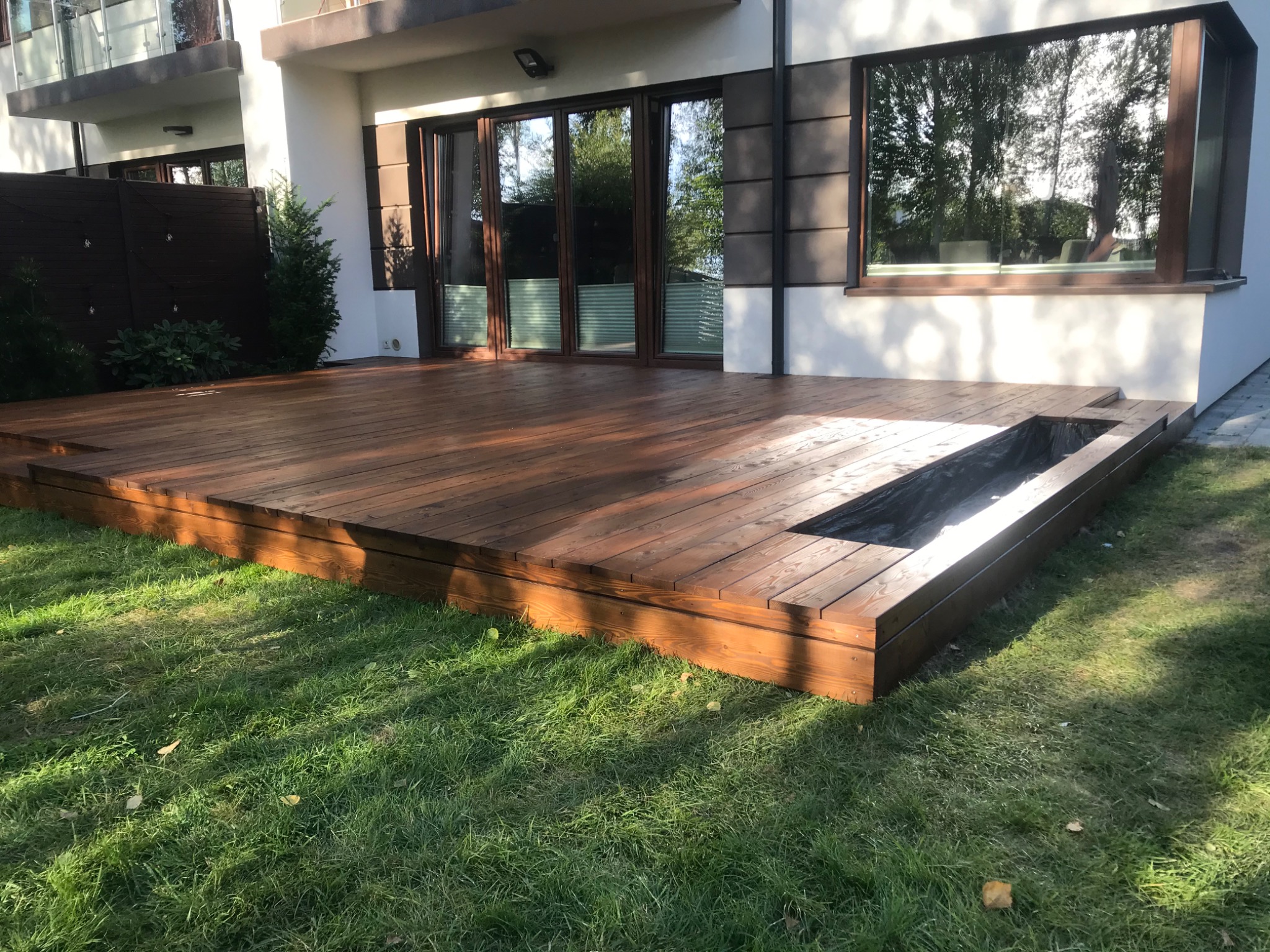 Nowoczesny, drewniany taras przylegający do domu z wbudowaną donicą, widok z poziomu trawnika w słoneczny dzień.