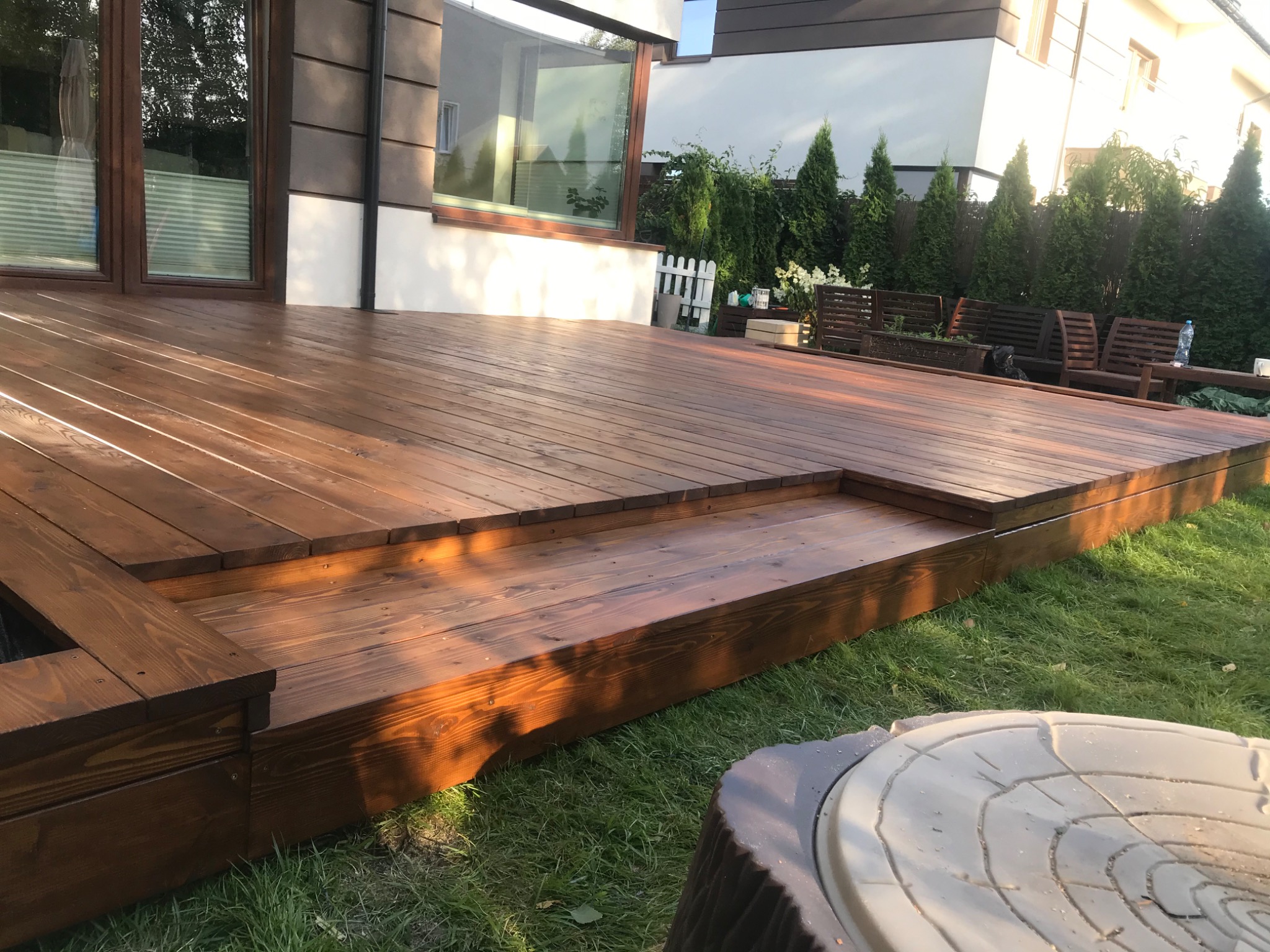 Nowoczesny taras drewniany z dwoma stopniami, wykonany z desek o ciepłym, brązowym odcieniu, przylegający do jasnego domu z dużymi oknami, otoczony zielonym trawnikiem i tujami.