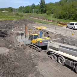 Żółta koparka Volvo nabiera ziemię do wywrotki na placu budowy, obok widoczne zabezpieczenia wykopu i linia kolejowa w tle.