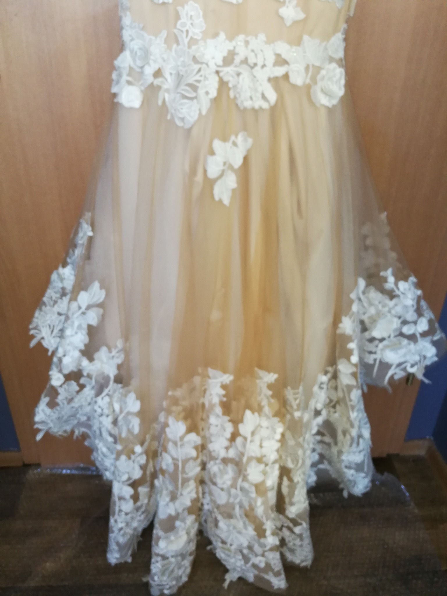 Beżowa suknia ślubna z białymi, koronkowymi aplikacjami kwiatowymi, widoczne detale wykończenia dołu sukni.