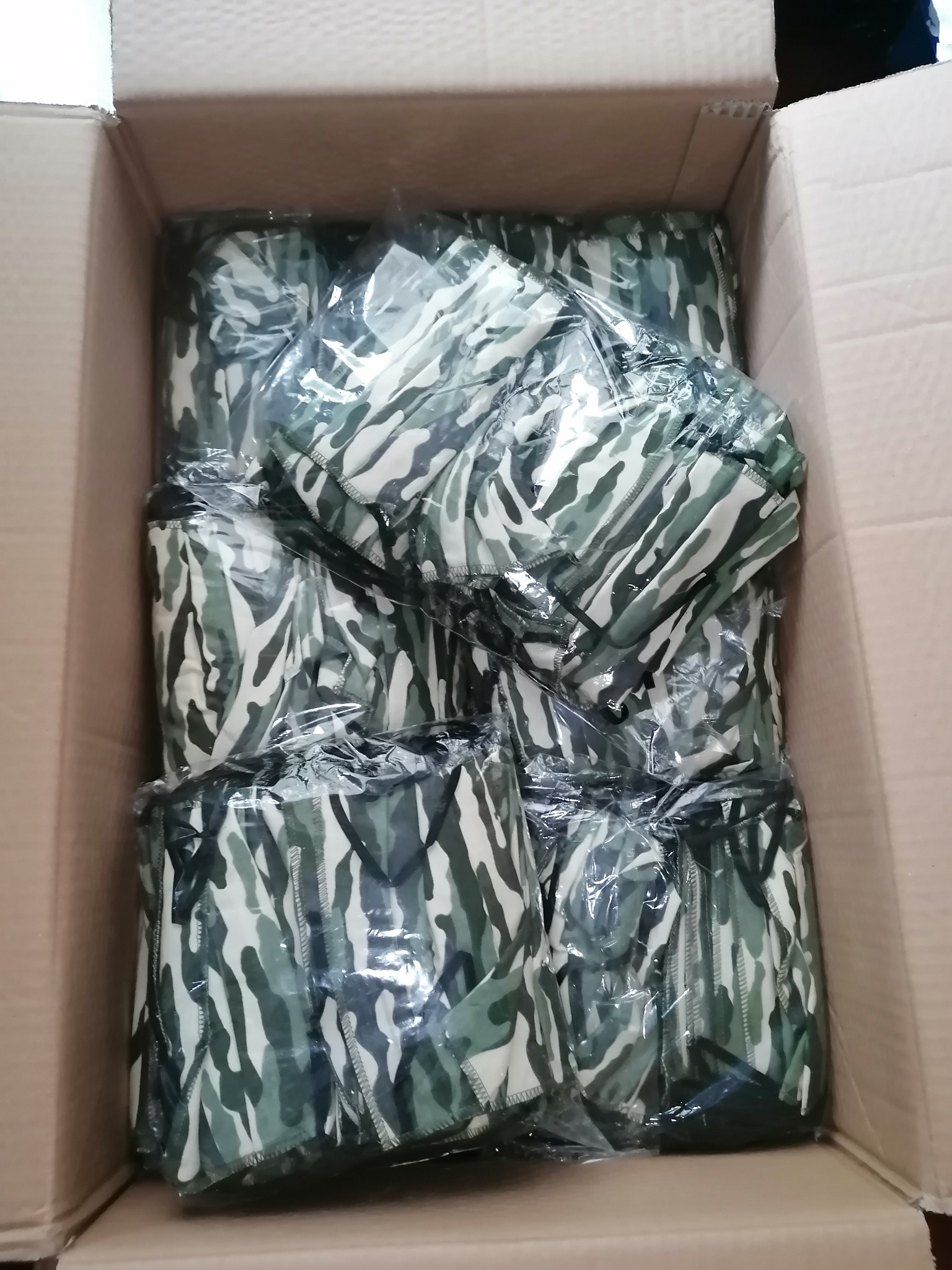 Partia maseczek ochronnych w kamuflażu, zapakowanych w folię i umieszczonych w kartonowym pudle, widok z góry.