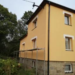 Dwukondygnacyjny dom z żółtą elewacją, białymi obramowaniami okien i kamienną podmurówką, częściowo zasłonięty rusztowaniem, otoczony zielenią.