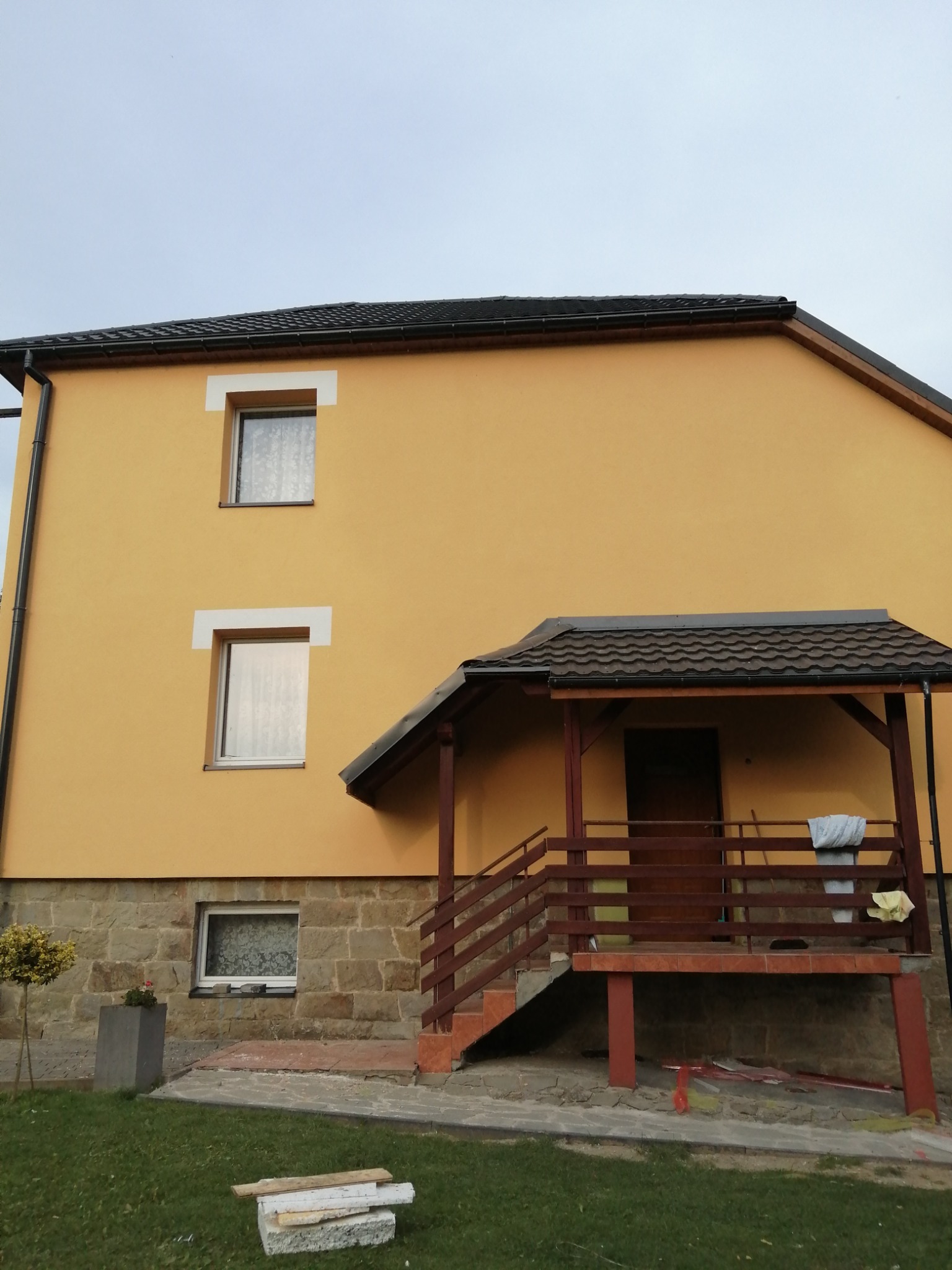 Dwupiętrowy dom z żółtą elewacją, kamienną podmurówką i drewnianym gankiem z brązową balustradą. Na trawniku leżą deski i kamienie.