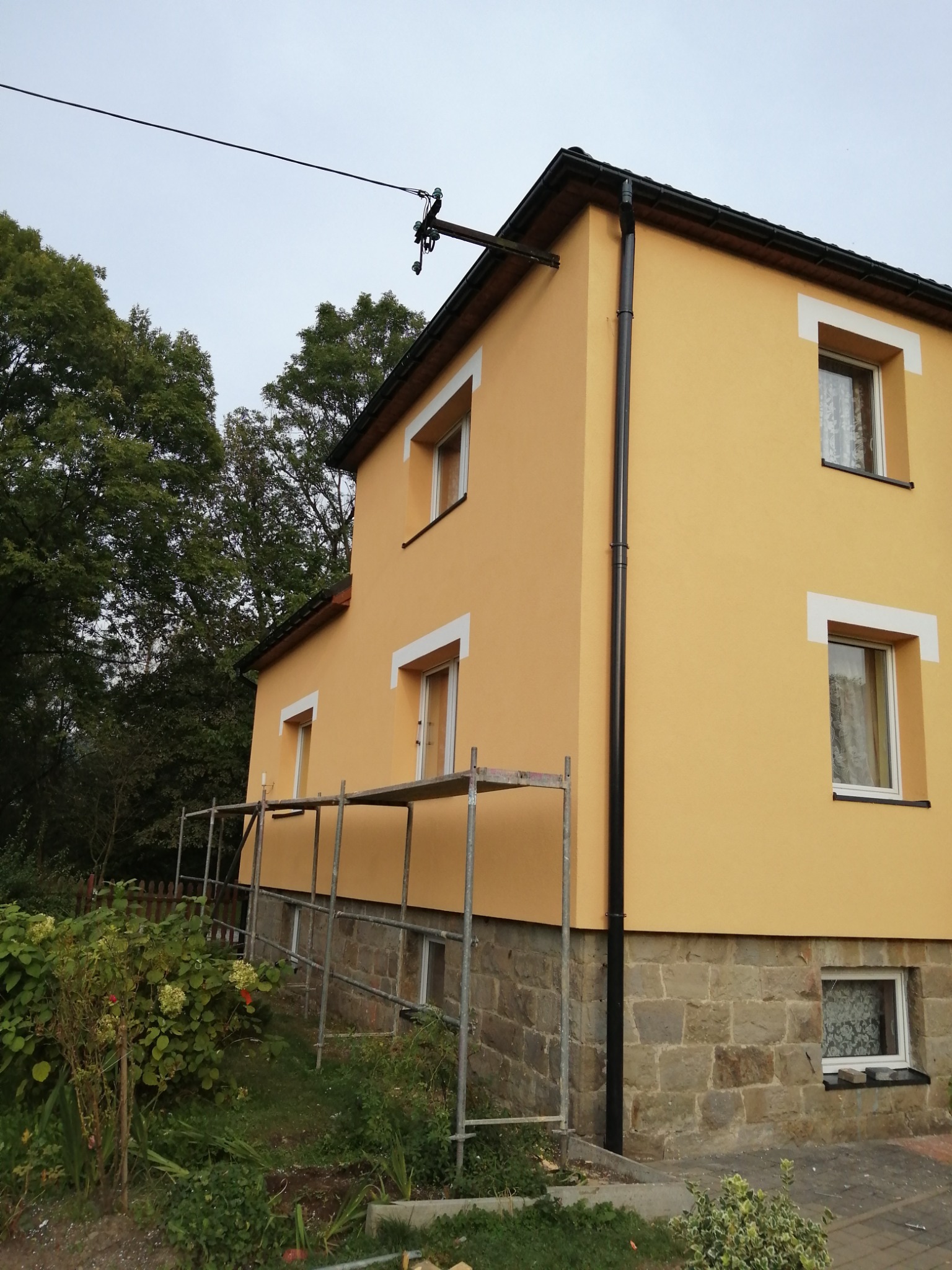 Dwukondygnacyjny dom z żółtą elewacją, białymi obramowaniami okien i kamienną podmurówką, częściowo zasłonięty rusztowaniem, otoczony zielenią.