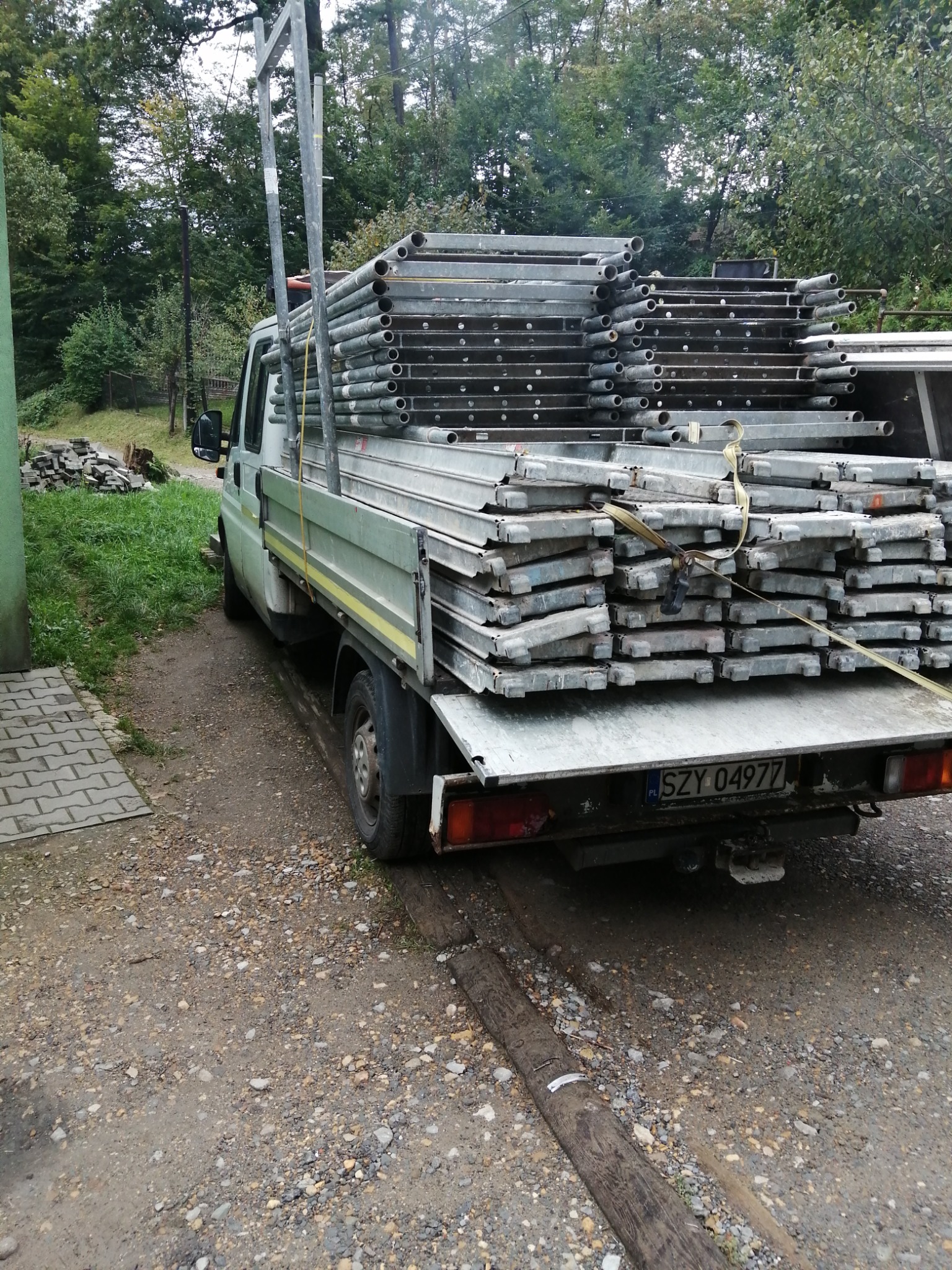 Ciężarówka z załadowanym rusztowaniem metalowym, zaparkowana na żwirowej drodze w pobliżu zieleni.