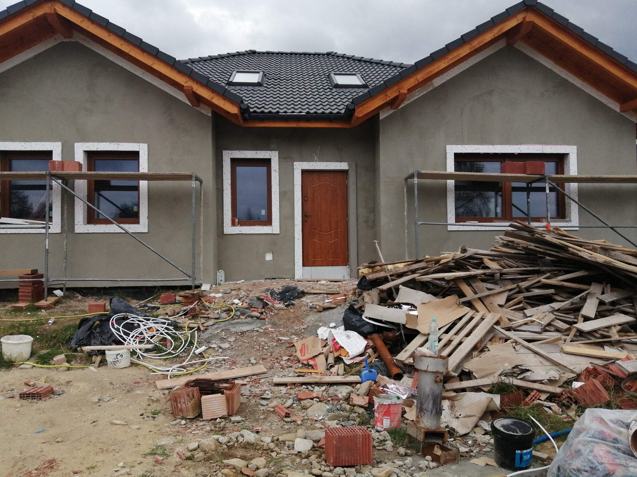 Dom jednorodzinny w trakcie prac elewacyjnych: surowa, szara elewacja, drewniana stolarka okienna i drzwiowa z białymi obramowaniami, rusztowania, stos gruzu i odpadów budowlanych przed budynkiem.