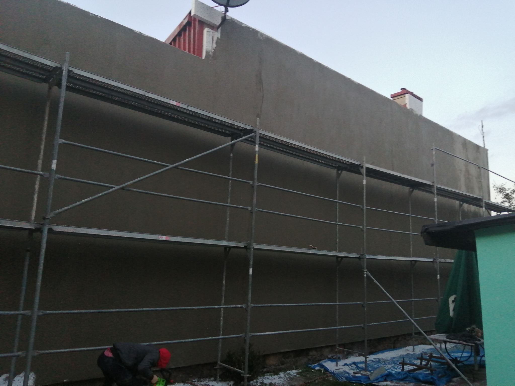 Świeżo nałożony, szary tynk na ścianie budynku, widoczne rusztowanie i pracownik w czerwonej czapce przy ziemi, fragment dachu sąsiedniego budynku w tle.