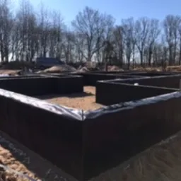 Fundamenty domu jednorodzinnego w trakcie izolacji przeciwwilgociowej, widoczna czarna folia ochronna na szalunku, w tle drzewa i zaparkowany van.