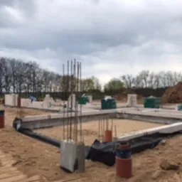 Fundamenty domu w trakcie budowy, widoczne szalunki, zbrojenie i bloczki fundamentowe. Otoczenie to plac budowy z piaskiem i ziemią, w tle drzewa.