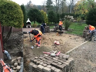 Prace brukarskie w ogrodzie: robotnicy w odblaskowych ubraniach układają kostkę brukową na przygotowanym podłożu, widoczne narzędzia, taczka i zaplecze ogrodowe.