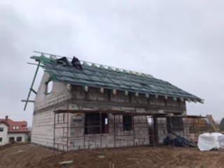 Budowa domu jednorodzinnego z białych bloczków, widoczne rusztowania i fragment dachu z zieloną membraną, dwóch robotników na dachu, pochmurne niebo.