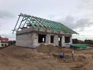 Surowy budynek z betonu komórkowego w trakcie budowy dachu, widoczne elementy konstrukcji drewnianej, hałda piasku przed budynkiem.