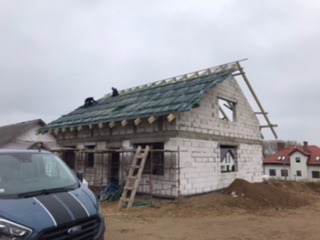 Budowa domu w trakcie: widok na niezadaszony budynek z białego pustaka, częściowo pokryty zieloną papą na dachu, drabina oparta o ścianę szczytową, w tle zabudowa jednorodzinna i błękitny samochód...