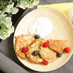 Fotografia produktowa: Naleśniki owsiane z jogurtem, malinami i borówkami na drewnianym talerzu, obok srebrne sztućce na żółtej serwetce i gałązka kwitnących białych kwiatów.