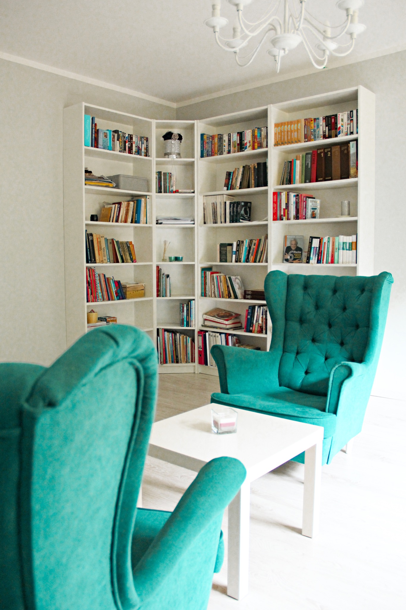 Jasne wnętrze z białymi regałami wypełnionymi książkami, turkusowymi fotelami typu uszak i białym stolikiem kawowym z zapaloną świecą.