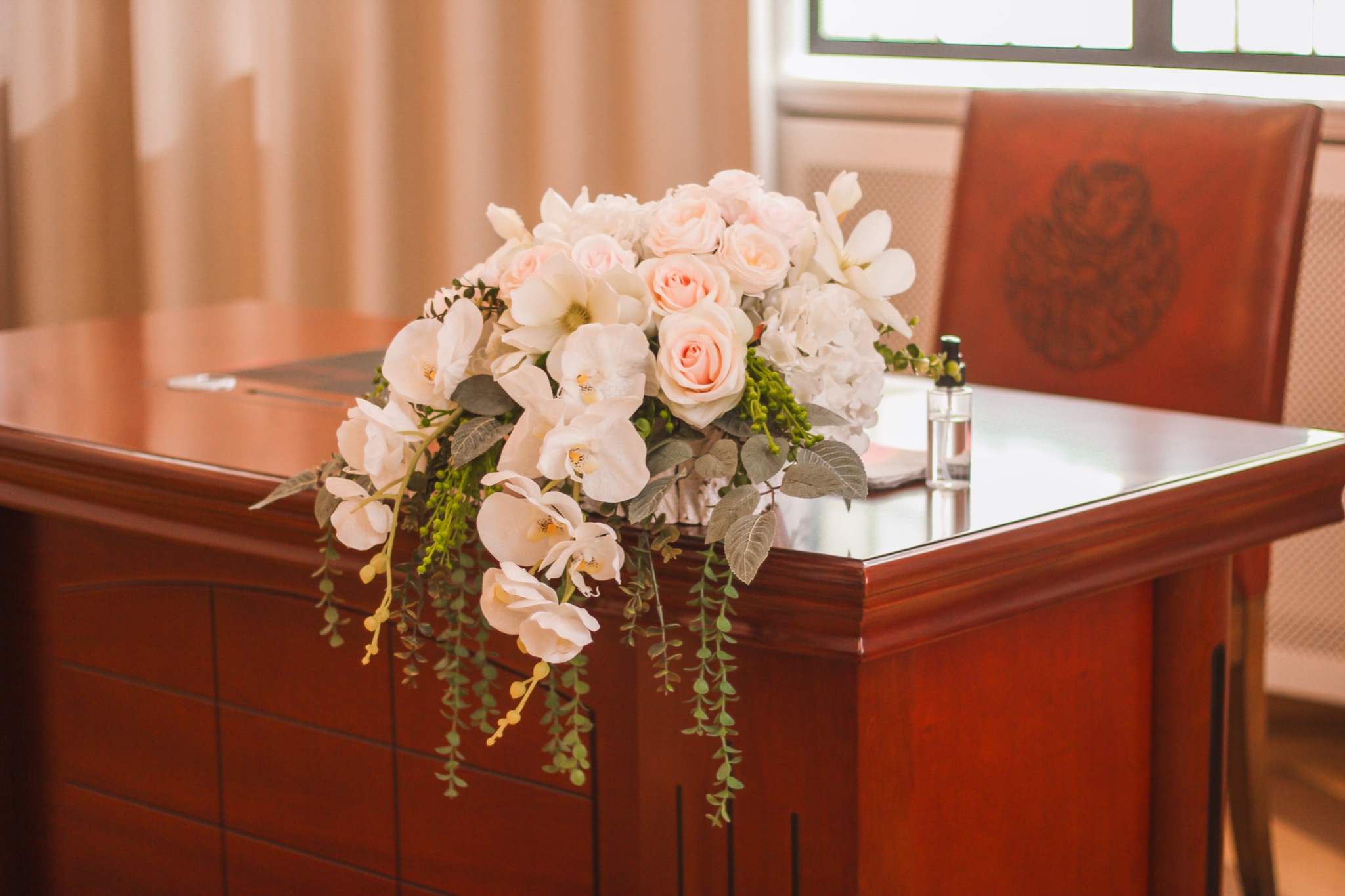 Dekoracja kwiatowa z białych orchidei, różowych róż i zielonych pnączy na mahoniowym stole z błyszczącym blatem, obok stoi flakonik perfum, w tle krzesło z herbem.