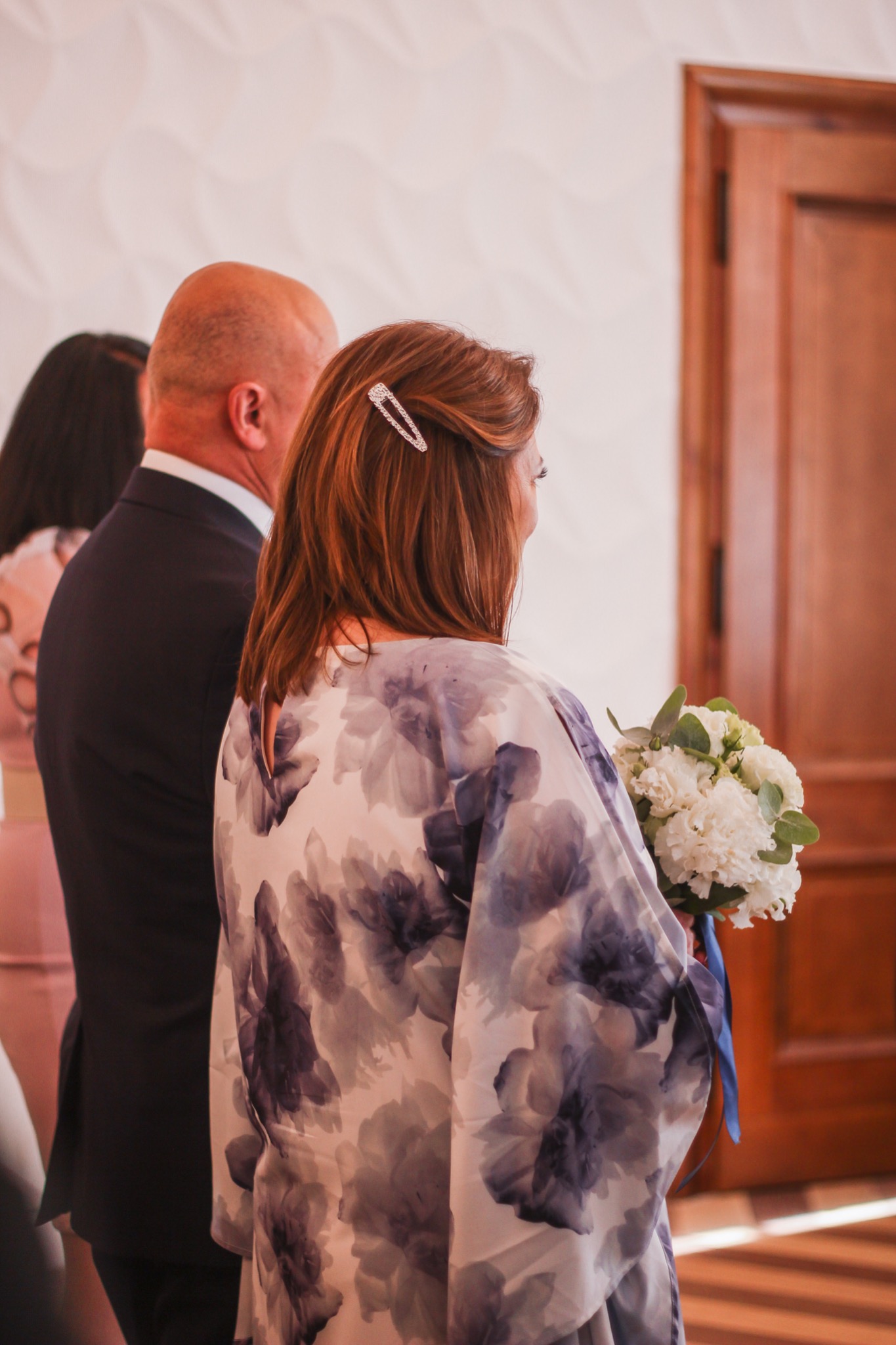 Kobieta z bukietem białych kwiatów, widziana od tyłu, z ozdobną spinką we włosach, w tle mężczyzna w garniturze i drewniane drzwi, Wrocław.