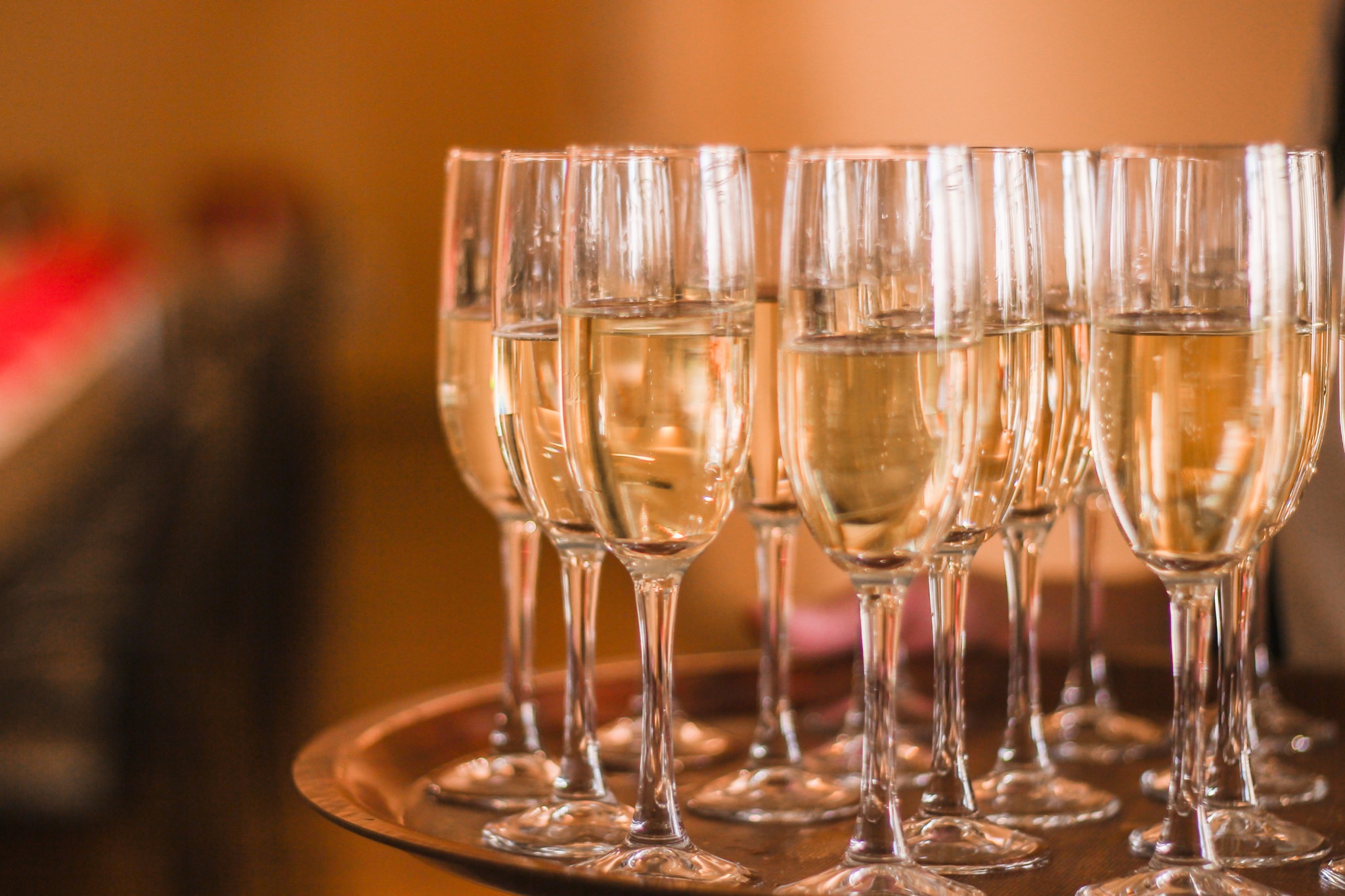 Rząd kieliszków do szampana wypełnionych napojem na drewnianej tacy, z delikatnym oświetleniem i rozmytym tłem.
