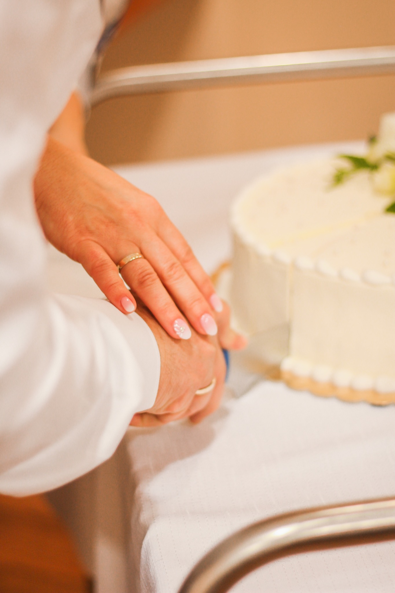 Dłonie pary młodej kroją tort weselny; widoczny złoty pierścionek na palcu kobiety i delikatny manicure z motywem kwiatowym.