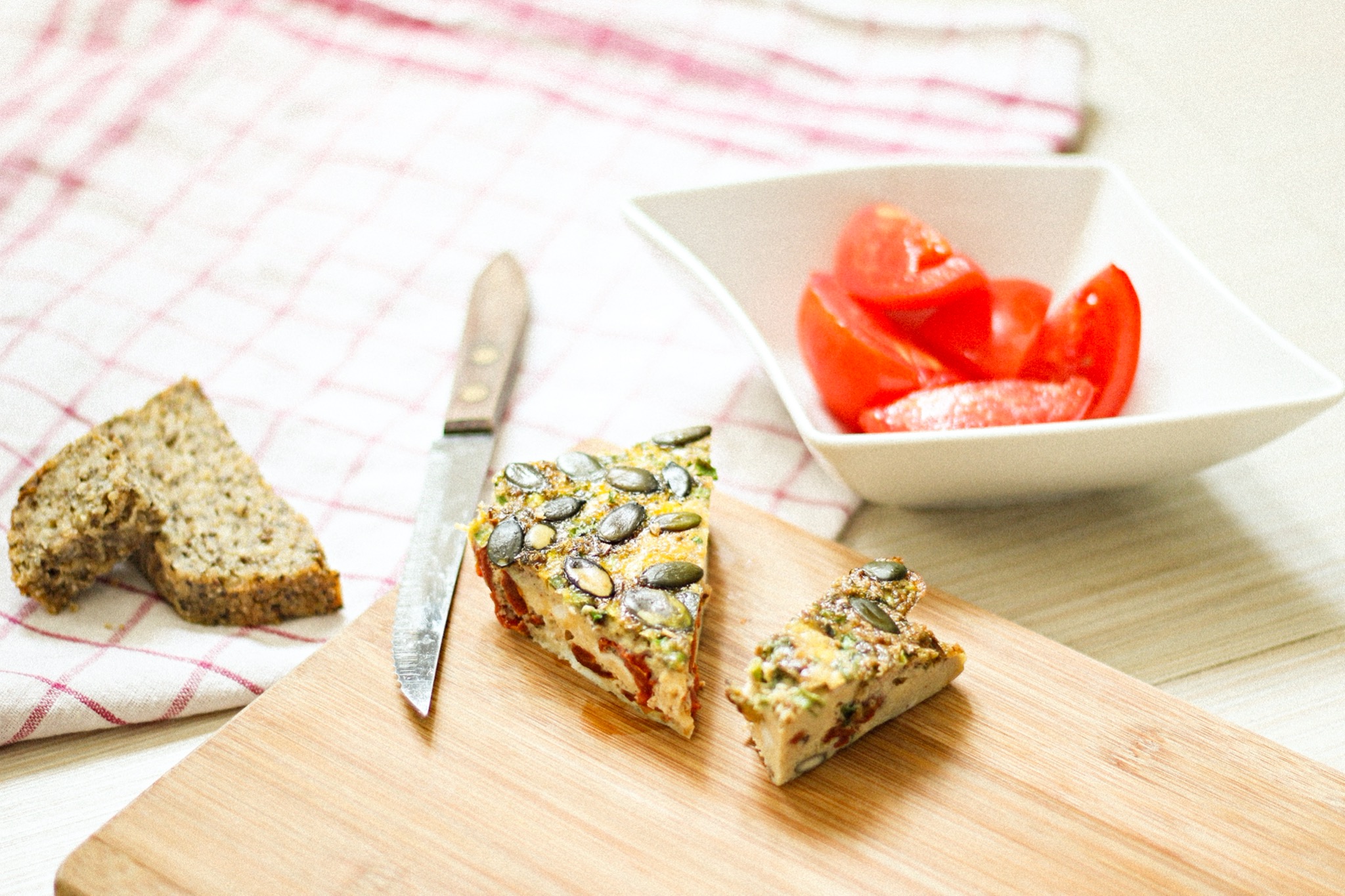 Kawałek quiche z pestkami dyni i suszonymi pomidorami na drewnianej desce, obok chleb razowy, nóż i miseczka z pokrojonymi pomidorami na tle kratowanej ściereczki.