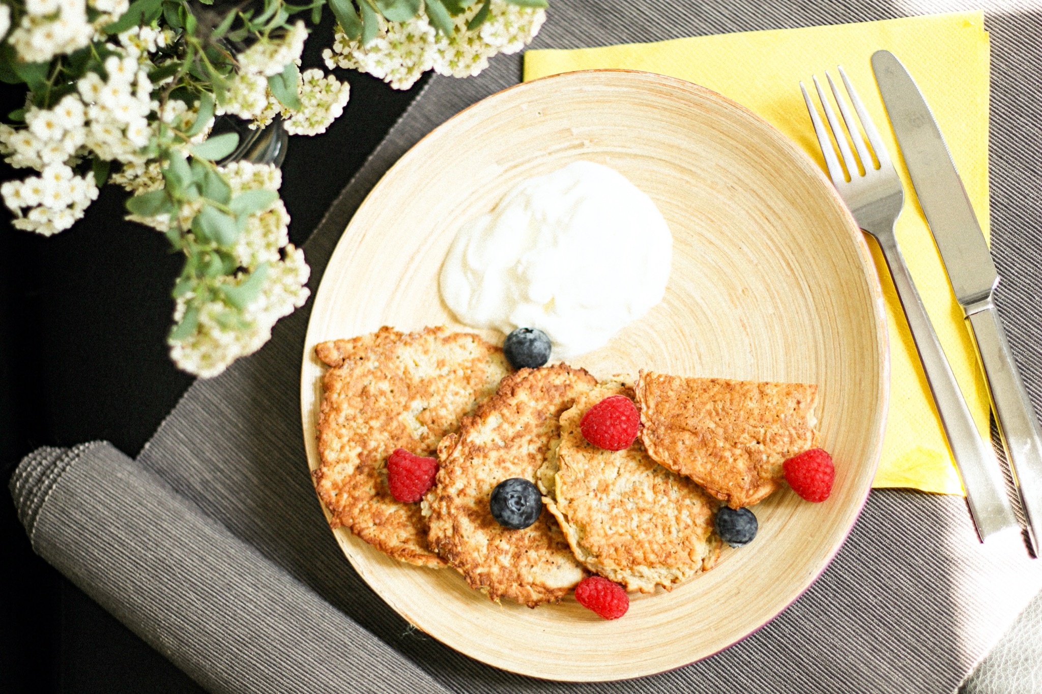 Fotografia produktowa: Naleśniki owsiane z jogurtem, malinami i borówkami na drewnianym talerzu, obok srebrne sztućce na żółtej serwetce i gałązka kwitnących białych kwiatów.