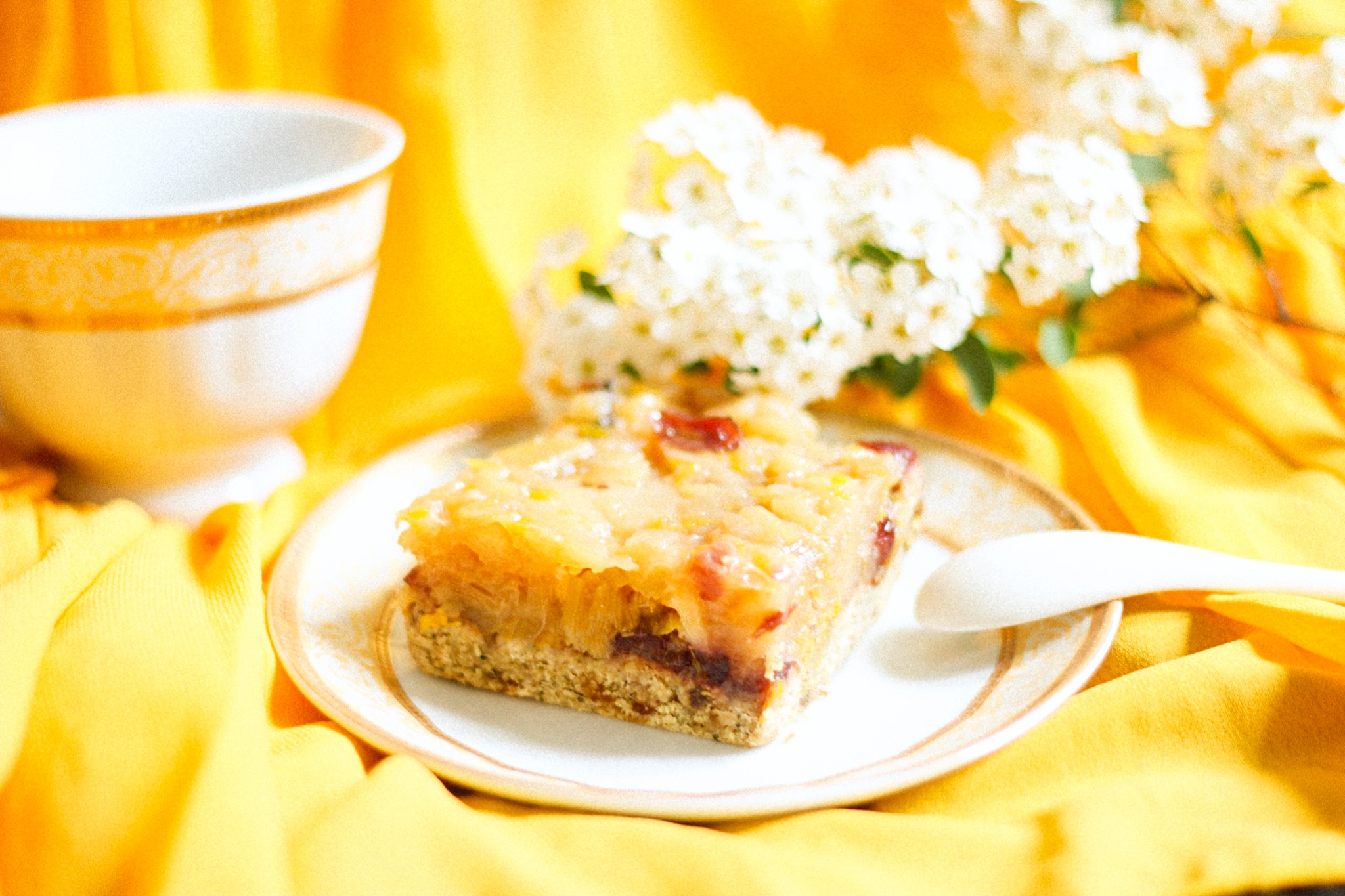Kwadratowy kawałek ciasta z owocami na talerzyku z łyżeczką, obok filiżanka i gałązka kwitnącego krzewu na żółtym tle
