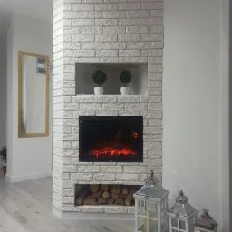 Wykończony narożny kominek z białej cegły dekoracyjnej, z wkładem elektrycznym imitującym ogień, półką na drewno i dekoracyjnymi lampionami na podłodze z paneli.