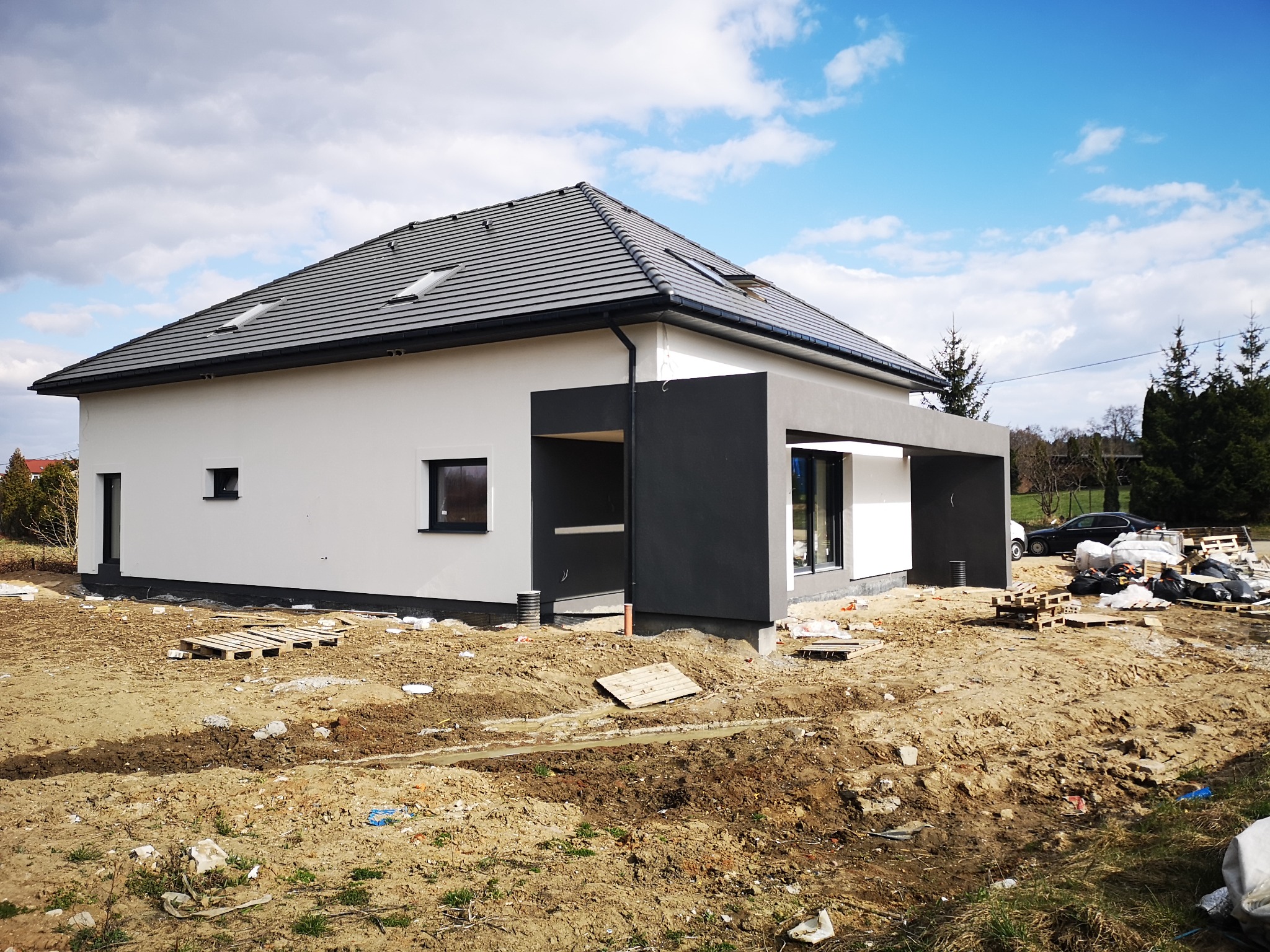 Nowoczesny dom jednorodzinny z płaskim dachem i oknami dachowymi, w trakcie prac wykończeniowych terenu wokół budynku, widoczne palety i worki z materiałami budowlanymi.