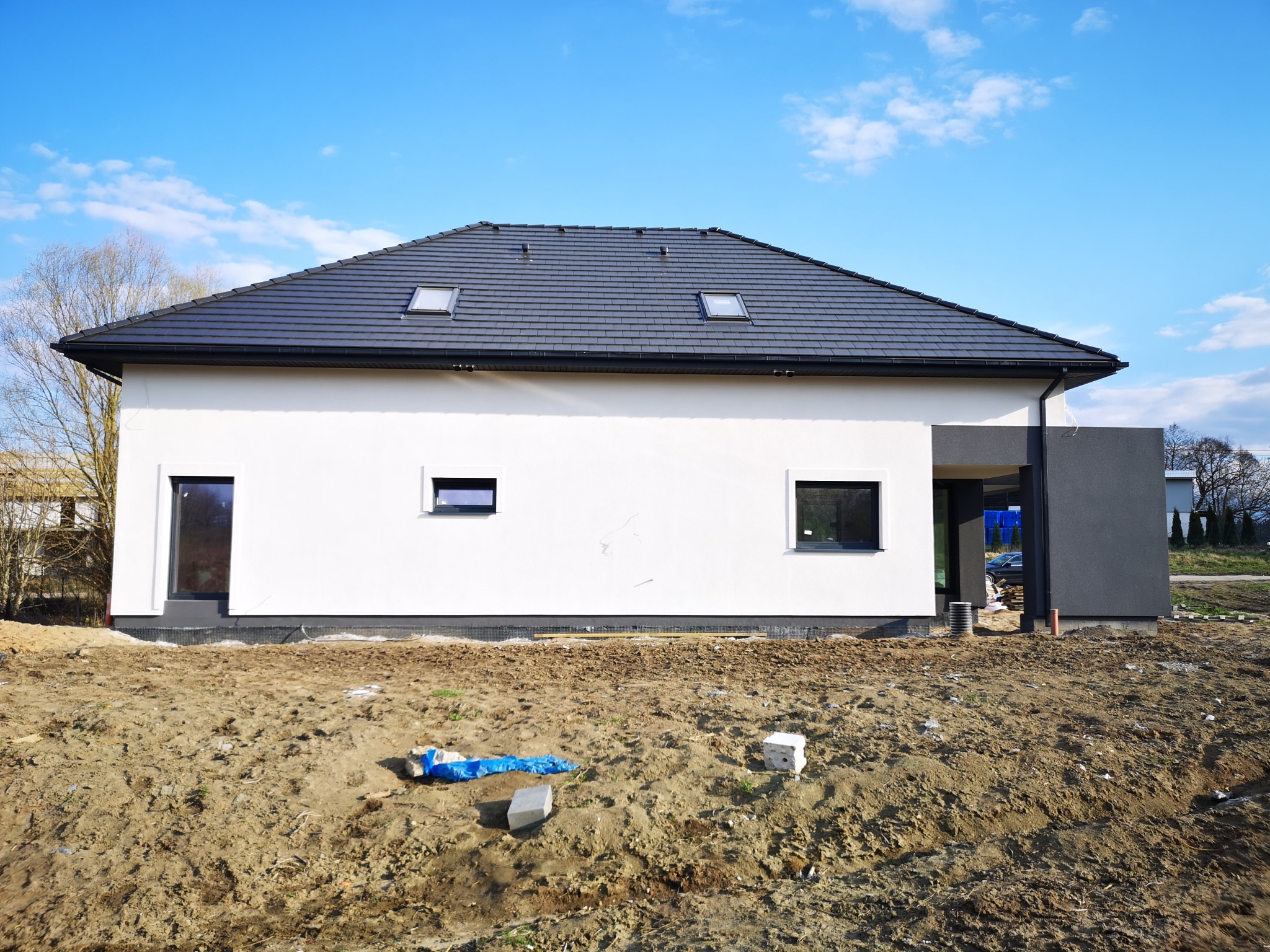 Nowoczesny dom jednorodzinny z białą elewacją i ciemnoszarą częścią, widoczne okna i dach z dachówką, teren wokół domu w trakcie prac ziemnych.