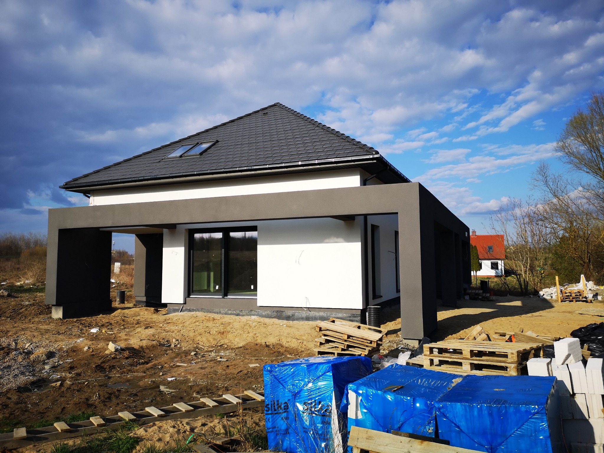 Nowoczesny dom jednorodzinny w trakcie budowy, z widoczną białą elewacją i szarą konstrukcją zadaszenia, na pierwszym planie zabezpieczone materiały budowlane w niebieskiej folii.