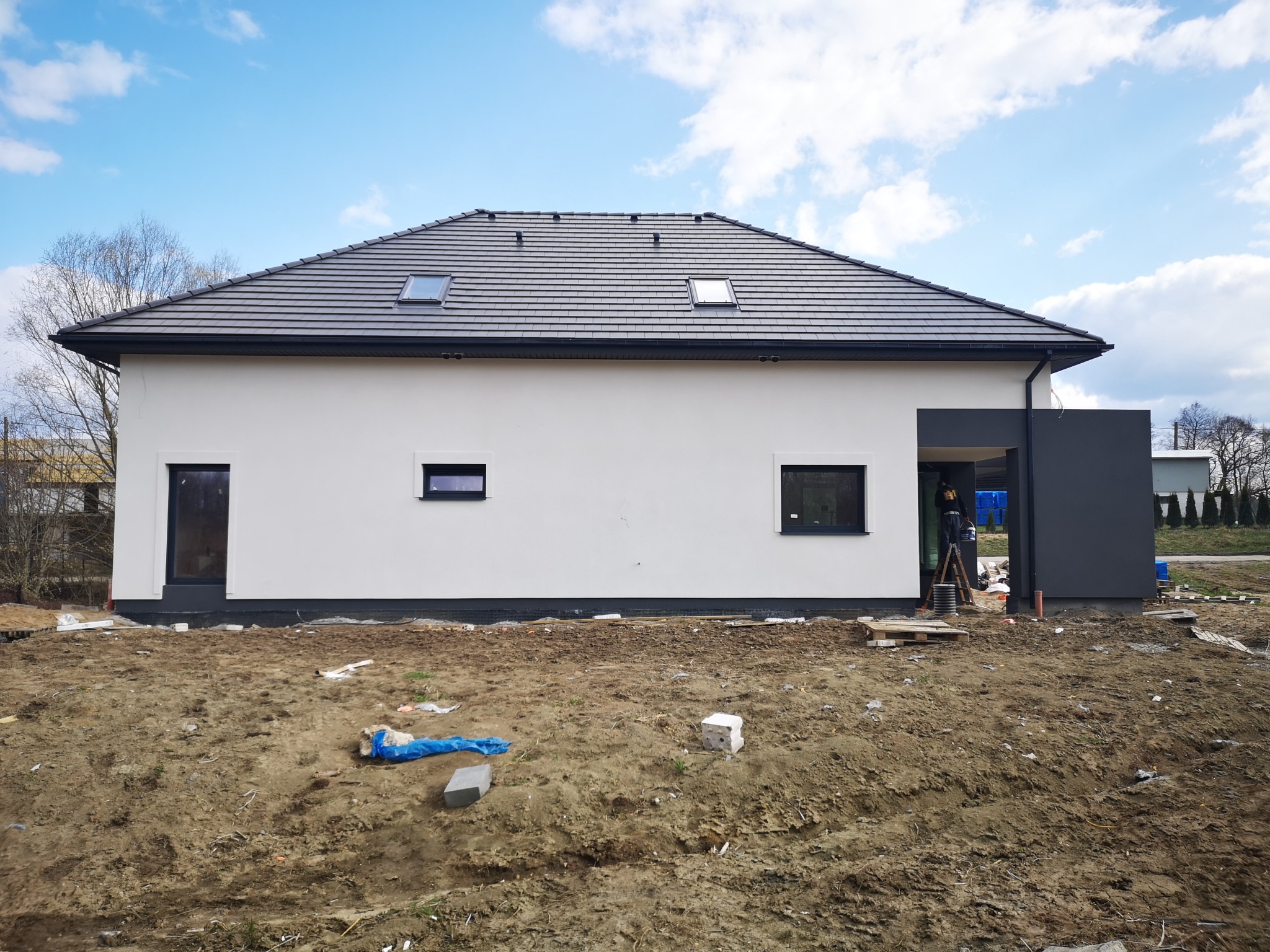 Nowoczesny dom jednorodzinny z białymi ścianami i ciemną dachówką, widok z zewnątrz na niezagospodarowany teren budowy, z widocznymi elementami wykończeniowymi i oknami dachowymi.