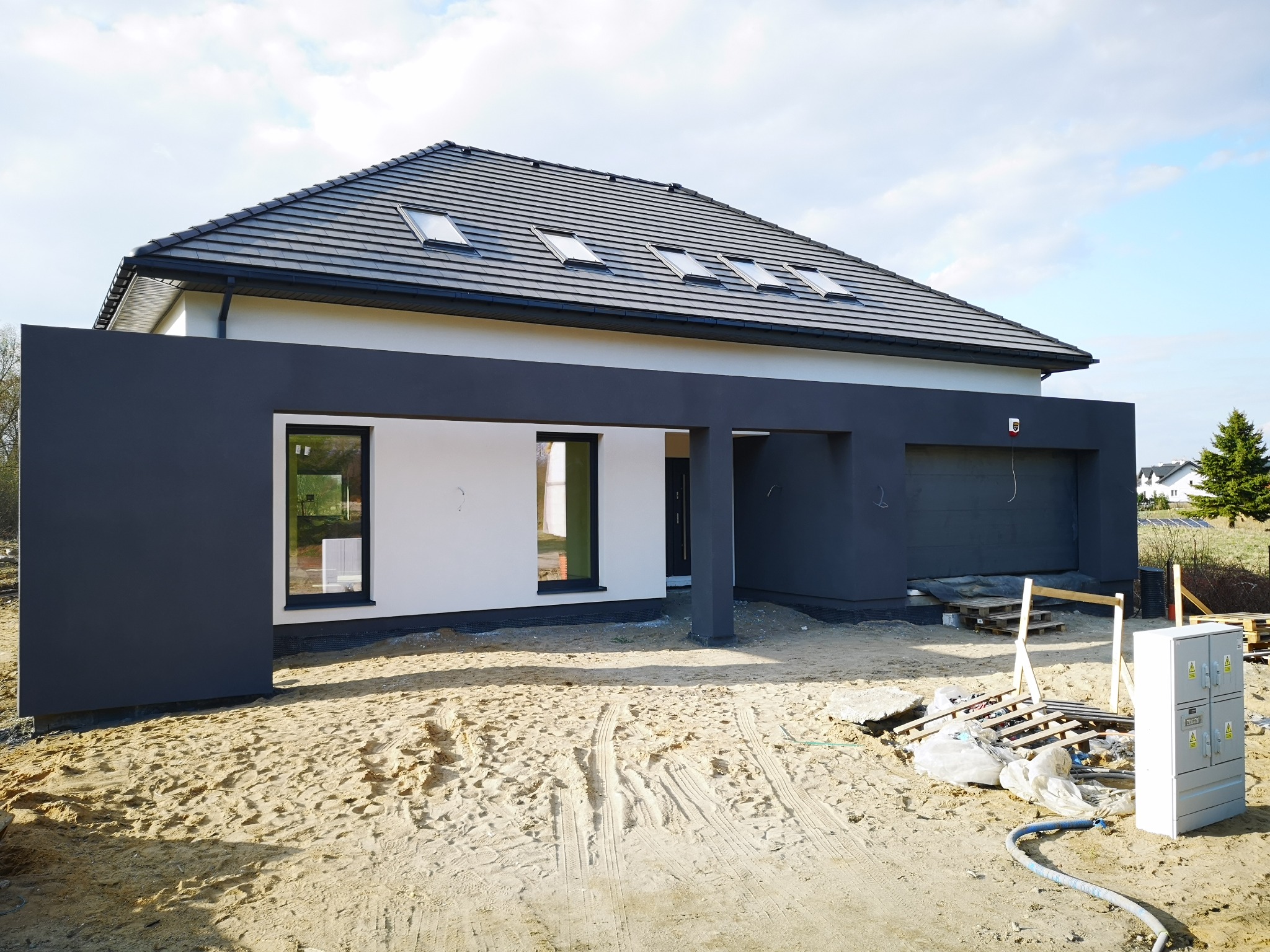 Nowoczesny dom jednorodzinny z płaskim dachem i oknami dachowymi, część elewacji w kolorze grafitowym, teren wokół w trakcie prac wykończeniowych, widoczne piasek, materiały budowlane i skrzynka...