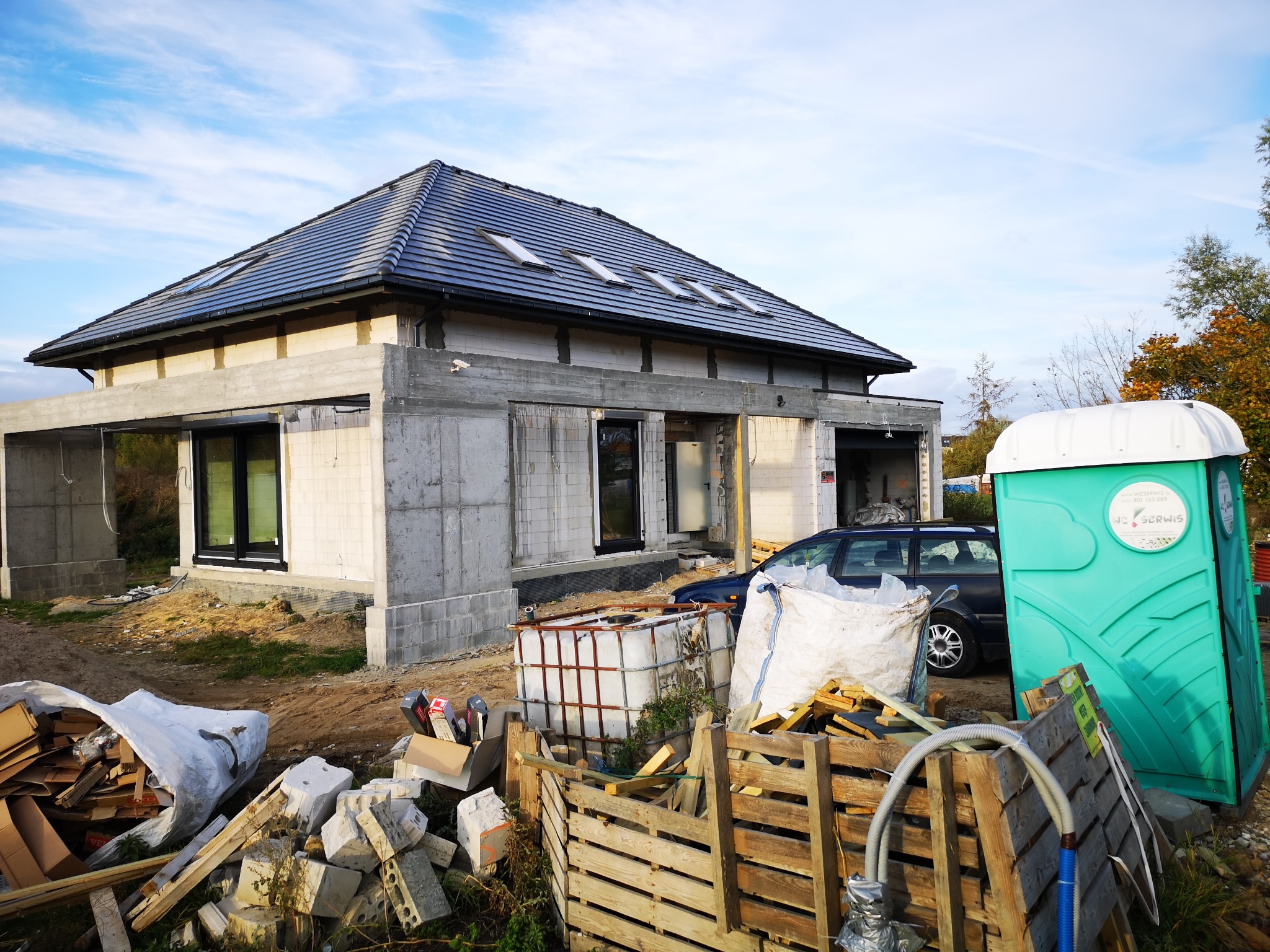 Surowy stan domu jednorodzinnego w trakcie budowy z widocznymi pustakami, betonowymi nadprożami, oknami PCV, dachem pokrytym dachówką oraz tymczasową toaletą przenośną na placu budowy.