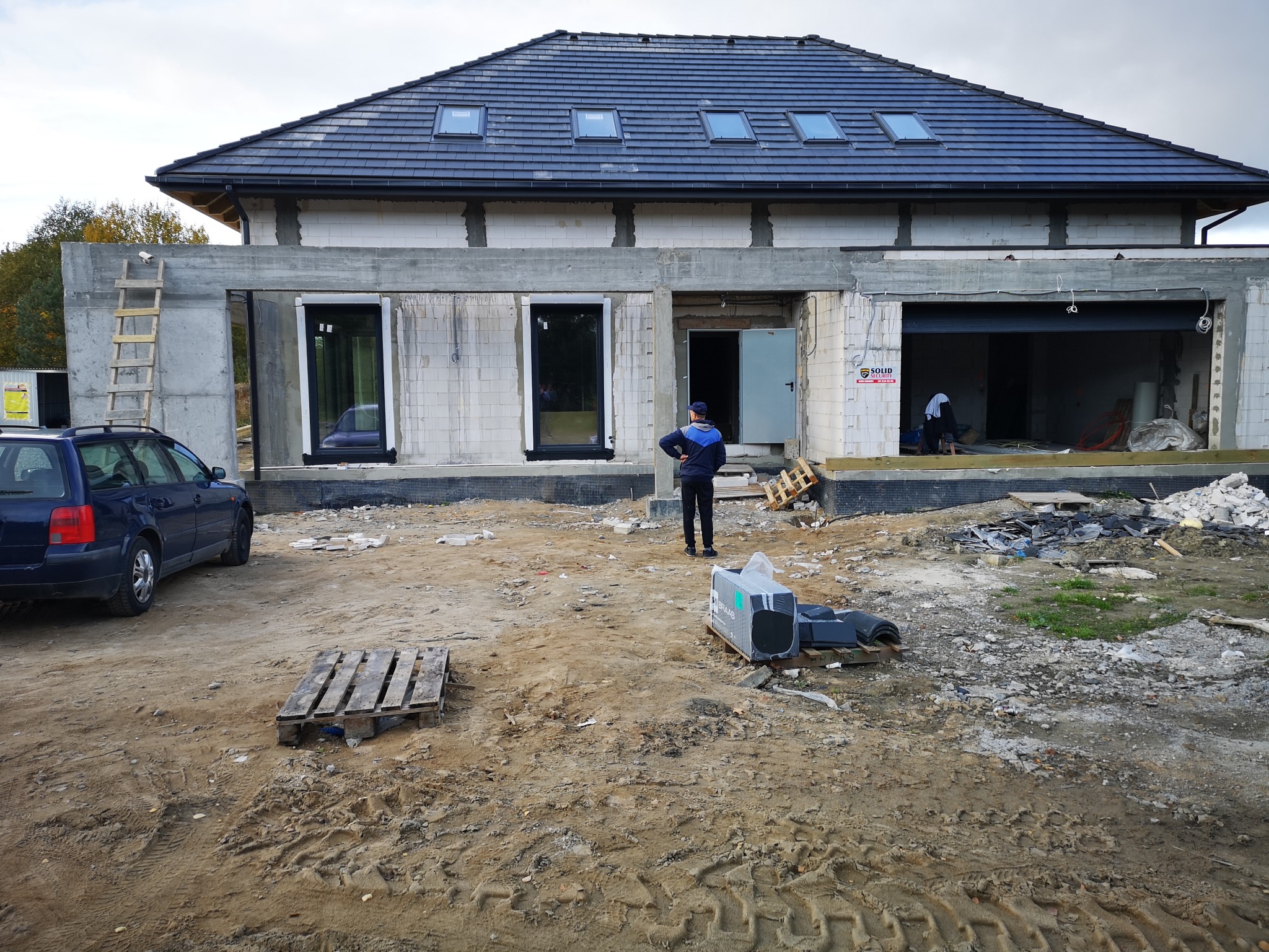 Niedokończony dom jednorodzinny z ciemną dachówką i oknami dachowymi, widoczny pracownik w kurtce i czapce, niezagospodarowany teren budowy z gruzem i paletami, niebieski samochód osobowy...