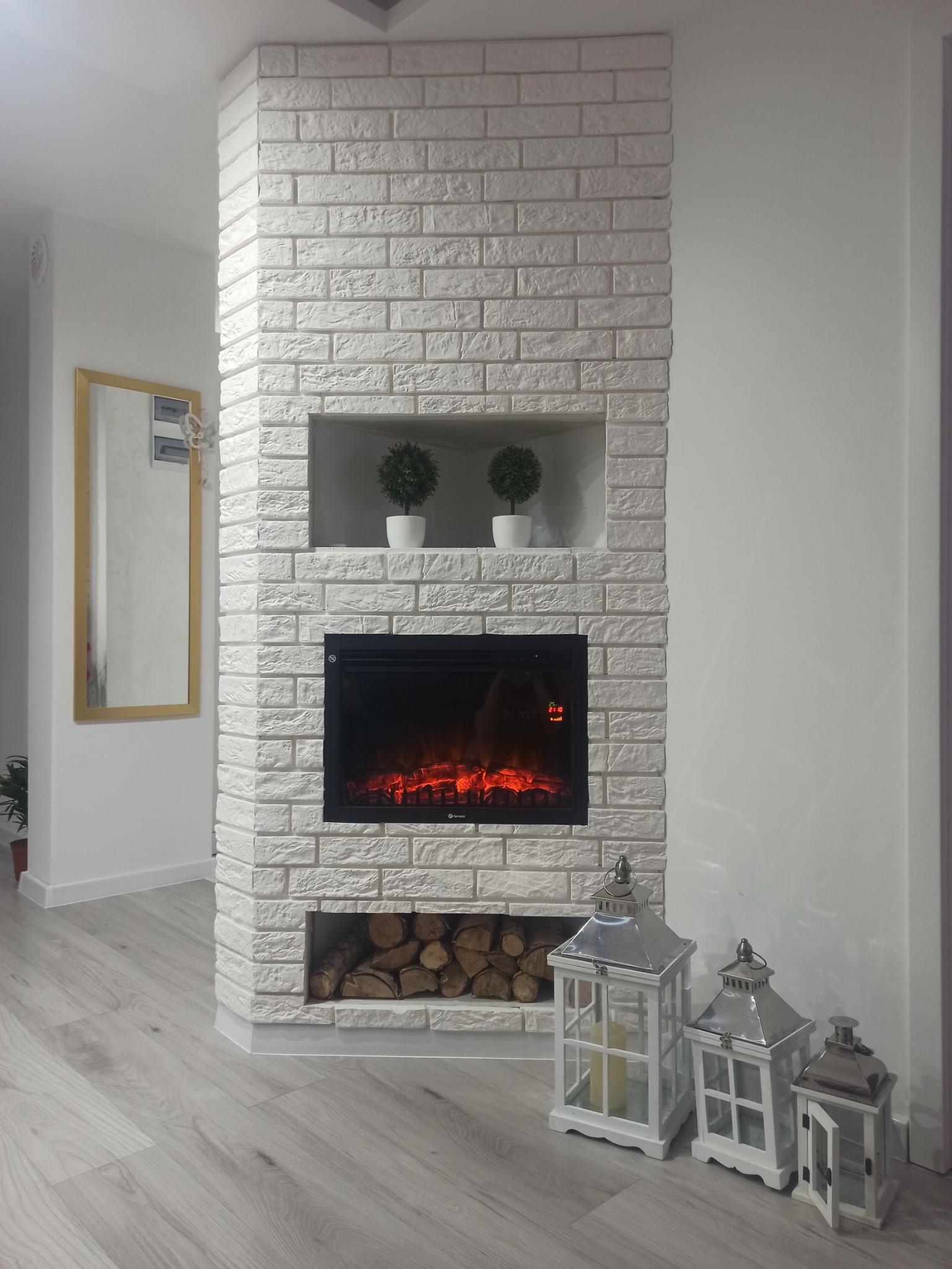 Wykończony narożny kominek z białej cegły dekoracyjnej, z wkładem elektrycznym imitującym ogień, półką na drewno i dekoracyjnymi lampionami na podłodze z paneli.